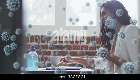 Image of covid 19 cells over woman in face mask, using laptop, drinking coffee in office Stock Photo