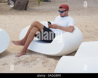 Man in a white t-shirt and sunglasses sits comfortably on white lounge chair at the beach using smartphone. Stock Photo