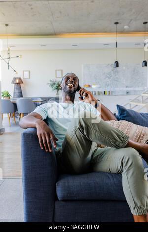 African American man relaxing on sofa, laughing while talking on smartphone. Communication, cellphone, lifestyle, leisure, happiness, relaxation Stock Photo