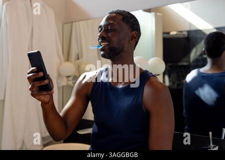 African American African American man brushing teeth while smiling at smartphone in bathroom. Hygiene, grooming, technology, morning routine, self-car Stock Photo