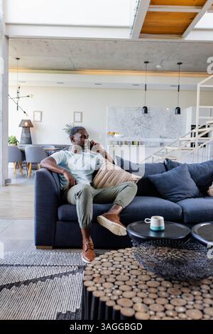 African American man sitting on sofa at home, talking on smartphone, looking relaxed. Conversation, communication, technology, relaxation, domestic, l Stock Photo