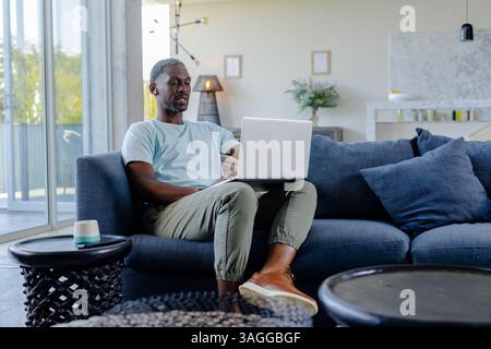 African American African American man using laptop on sofa at home, working remotely. Remote working, telecommuting, freelancer, technology, digital n Stock Photo