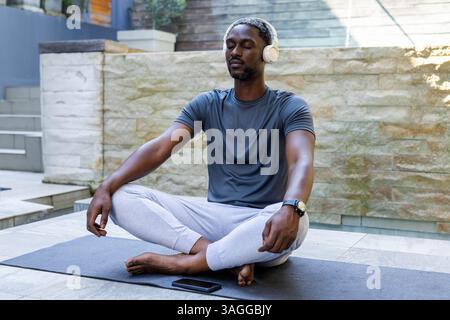 Meditating by pool, African American man in headphones relaxing on yoga mat, eyes closed. Relaxation, mindfulness, wellness, tranquility, meditation, Stock Photo