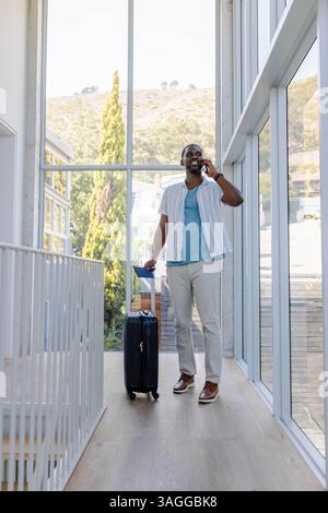 African American African American man talking on smartphone while pulling suitcase in modern hallway. Business, travel, professional, communication, t Stock Photo
