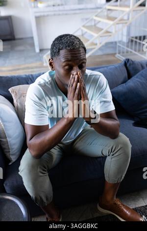 African American man sitting on couch at home, deep in thought with hands clasped. Contemplation, relaxation, solitude, introspection, mindfulness, tr Stock Photo