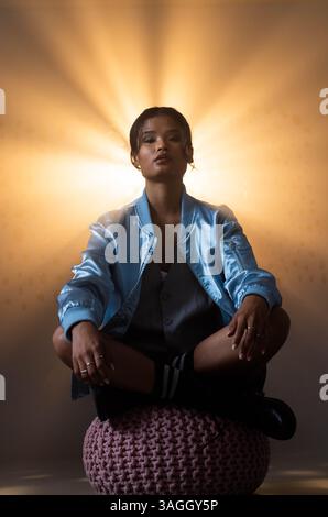 Sitting cross-legged on pouf, woman in blue jacket under dramatic light. Portrait, fashion, stylish, trendy, contemporary, dramatic lighting Stock Photo