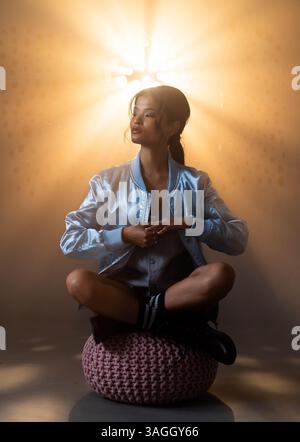 Young woman in bomber jacket sitting cross-legged on pouf, illuminated by light. Fashion, casual, style, cozy, relaxed, modern Stock Photo