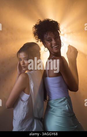 Posing back-to-back, two women smiling confidently in dramatic lighting setup. portrait, studio, friendship, empowerment Stock Photo