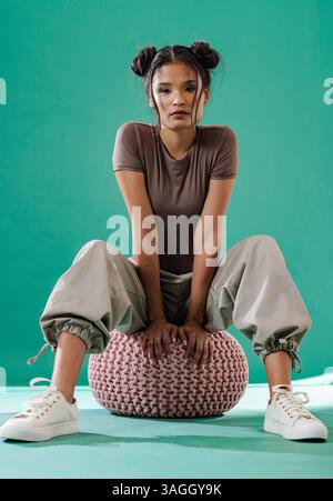 Asian woman in casual outfit sitting on pouf, looking confidently at camera. Confidence, portrait, fashion, stylish, poise, indoors Stock Photo