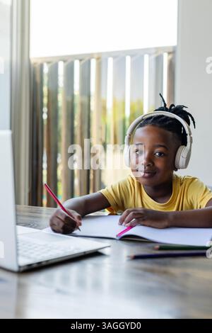 African American boy wearing headphones writing in notebook at home, using laptop for school. E-learning, homeschooling, digital, technology, studying Stock Photo