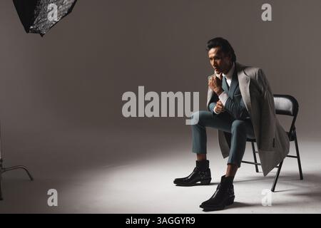 Stylish man in suit and coat sitting on chair, contemplating in studio. Fashion, style, business, contemplation, elegance, menswear Stock Photo