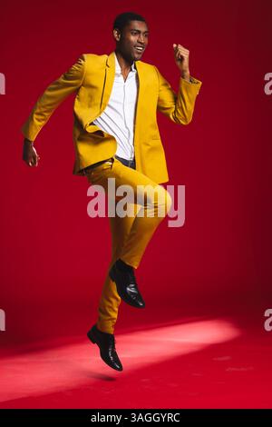 Man in vibrant yellow suit joyfully jumping against red studio backdrop. Fashion, colorful, enthusiasm, energy, studio shot, dynamic Stock Photo
