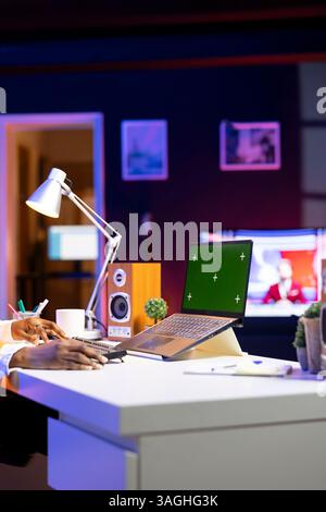 Person at home office desk using green screen mockup laptop, composing social media posts. Remote content creator employee monitoring engagement on chroma key isolated screen notebook Stock Photo