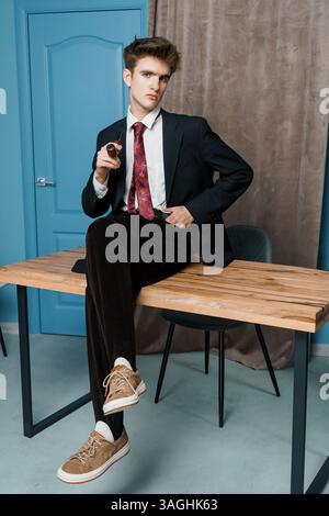 Stylish young man in a suit and sneakers casually sits on a wooden table, holding a pen with a confident expression against a mixed background. Stock Photo