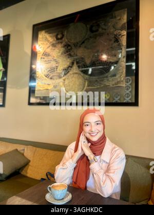 Smiling young woman in a headscarf enjoying coffee at a cafe table. Natural light, warm tones, relaxed atmosphere, indoor setting. Stock Photo