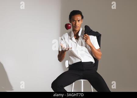Confident man in stylish outfit tossing apple while sitting on stool. Fashion, confidence, modern, casual, style, lifestyle Stock Photo