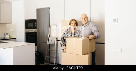 Happy senior couple smiling and holding cardboard boxes while relocating into new home Stock Photo