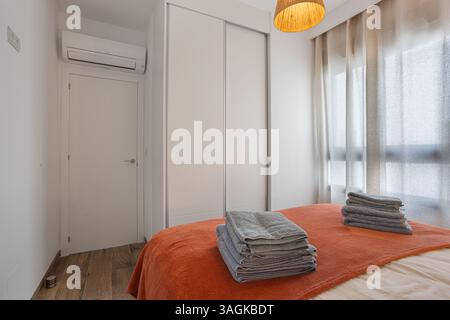 Side angle of a modern bedroom showcasing a coral bedspread, neatly folded grey towels, and minimalist built-in wardrobes. Soft filtered daylight enha Stock Photo