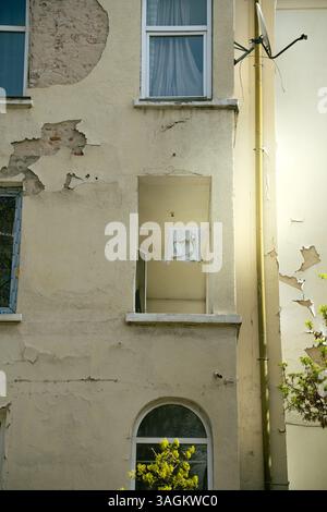 Urban apartment facade with peeling paint and damaged plaster, featuring broken balcony and sunlight falling between two old buildings. Stock Photo