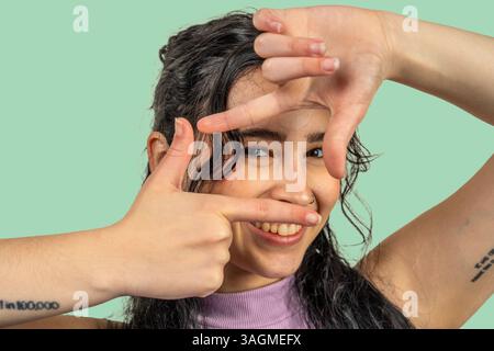 Cheerful young woman framing her face with fingers, smiling and enjoying the moment against a vibrant green background Stock Photo