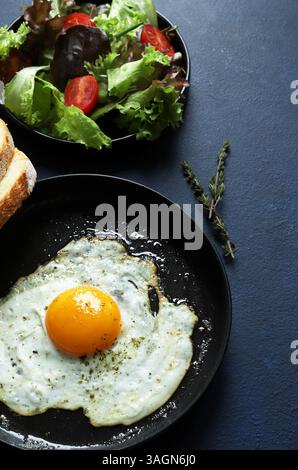 A single fried egg in a frying pan served with vegetables and bread on dark background. Healthy food concept. Directly above Stock Photo