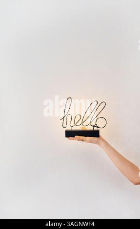 A woman's hand gently holds a decorative 'hello' sign with warm LED lights, creating a welcoming and minimalist scene. Stock Photo