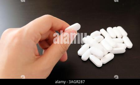 Hand picking up a white supplement tablet from a pile that is placed on a black table. Stock Photo