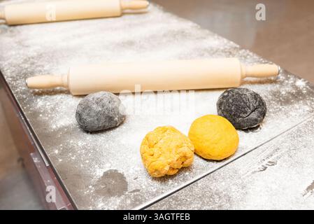 Assorted yellow and black pasta dough balls on a floured countertop with wooden rolling pins, ready for cooking or baking. Stock Photo
