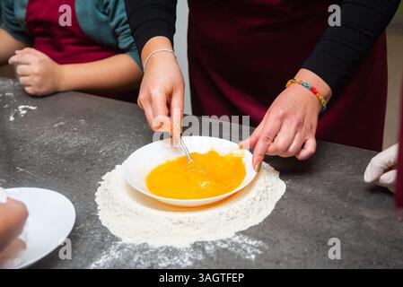 Close-up of hands beating egg yolks in a white bowl surrounded by flour on a kitchen counter, preparing fresh pasta dough. Stock Photo