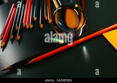 Creative still life composition with a clear glass ball reflecting colorful pencils on a dark green surface with artistic lighting. Stock Photo