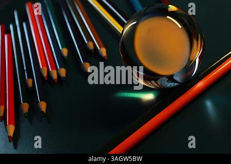 A creative macro shot using a clear glass ball placed among several sharpened colored pencils on a dark surface. Stock Photo