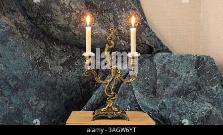 Vintage ornate brass candelabra with two burning white candles against a textured blue and grey fabric background. Stock Photo