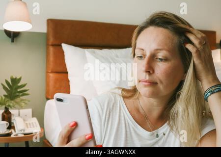 Beautiful blonde woman using smartphone lying on bed in bedroom Stock Photo