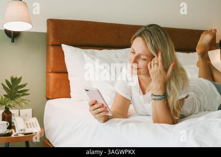 Beautiful blonde woman using smartphone lying on bed in bedroom Stock Photo