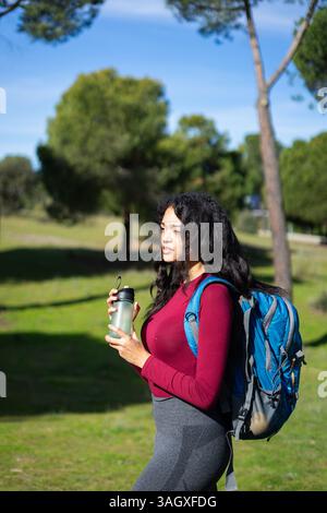 Young latin woman holding reusable water bottle, wearing backpack, enjoying break in nature Stock Photo