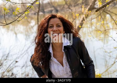 portrait of smiling and charming woman with red hair wearing leather jacket walking in nature Stock Photo