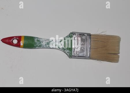 Old, worn-out paintbrush with green and red handle on a white background. Bristles are slightly splayed and the brush shows signs of paint stains and Stock Photo