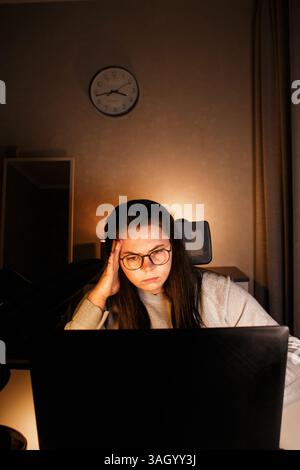 Young woman with eyeglasses working on laptop at home late at night, using modern technologies and getting online education. Remote learning concept. Stock Photo