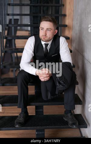 Man in a suit and vest sits on a set of stairs. He is wearing a tie and has a briefcase in his hand Stock Photo