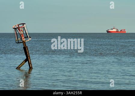 Weathered, rusted metal navigation beacon post leaning in calm coastal waters with a red offshore vessel on the distant horizon. Stock Photo