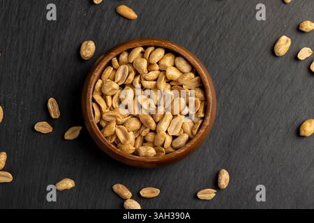 roasted peanuts in one wooden round bowl, crunchy peanuts without shells on a black slate board, top view Stock Photo