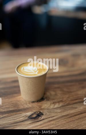A delicious latte with latte art in a disposable cup sits on a wooden table in a coffee shop. Stock Photo