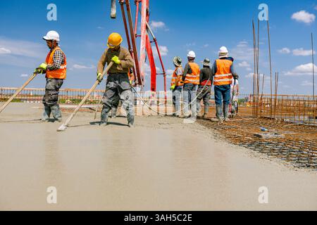Team of construction workers are pouring and leveling fresh concrete using handmade tool over rectangular armature. Stock Photo