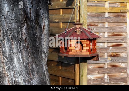 Wooden bird feeder made of wood in the form of a house hanging on a fence next to a tree. Stock Photo