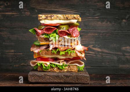 Extra large layered sandwich with ham, bacon, salami, cheese and fresh vegetables on dark wooden background.  Homemade food: big lunch mega sandwich. Stock Photo