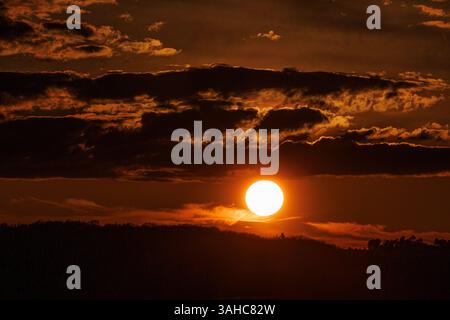The sun sets behind the rolling hills of Galicia, casting a fiery glow across the sky. Dark and orange clouds create a dramatic and moody atmosphere, Stock Photo