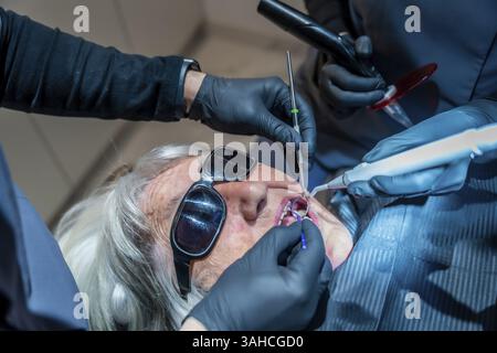 Dental professionals using specialized tools are performing a teeth whitening procedure on a senior woman patient wearing protective eyewear Stock Photo