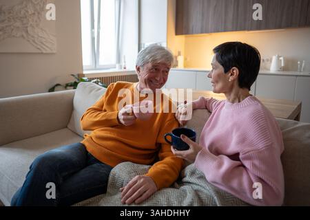 Happy engaged mature couple wife husband enjoys conversation while sipping tea together on cozy sofa Stock Photo