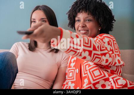 Two women smiling sofa watching TV together, enjoying time with friends at home Stock Photo