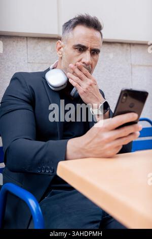 Man with headphones thinking while using phone and tablet at outdoor cafe table Stock Photo